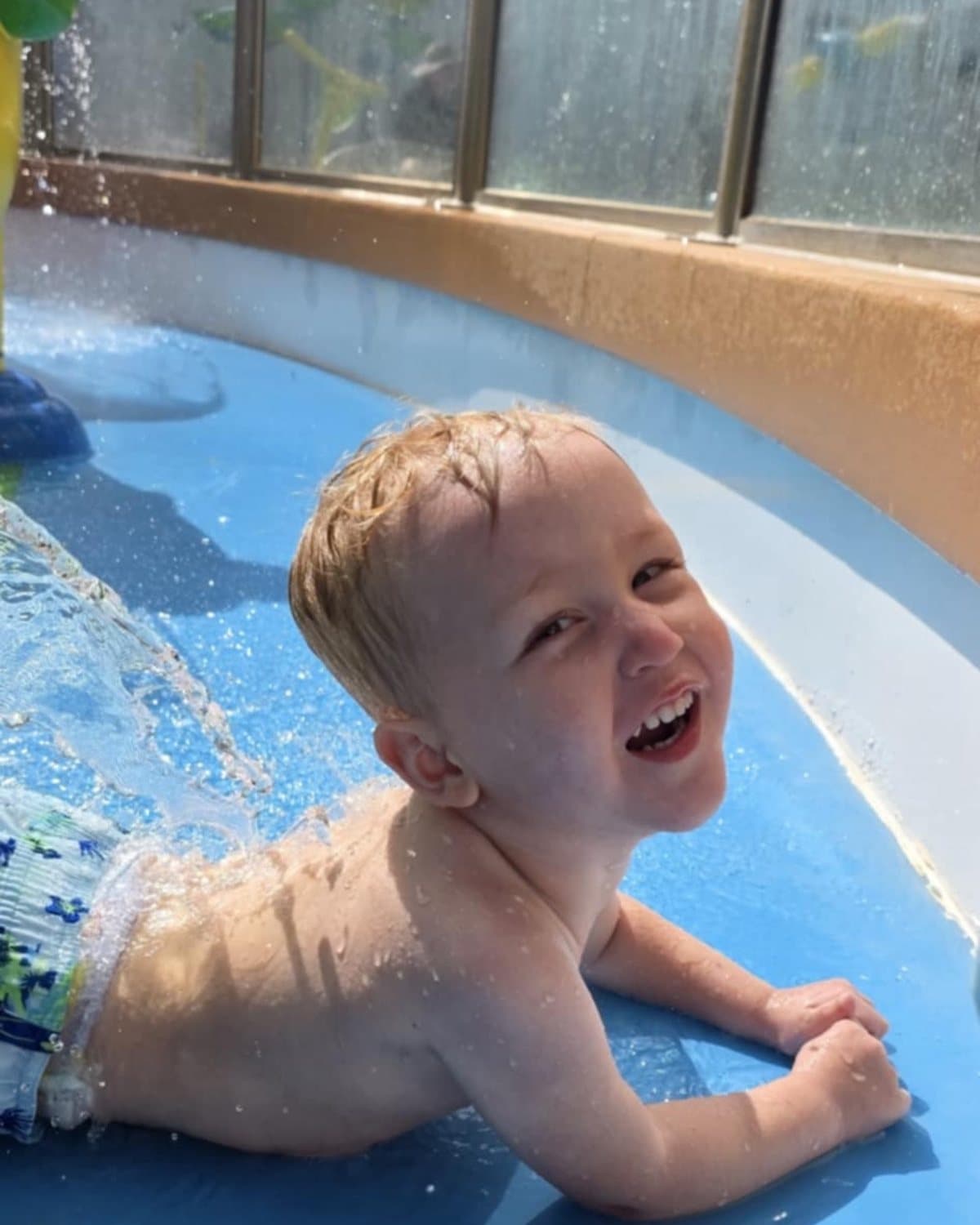 Maverick as a smiling toddler lying in the shallow water at a splash pad